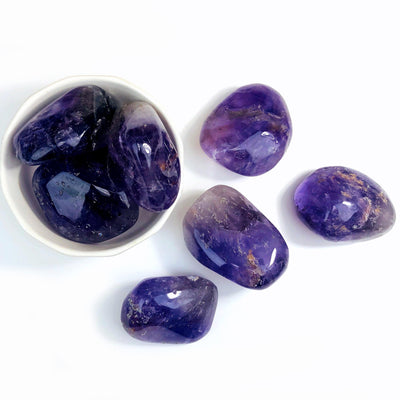bowl holding 3 amethyst tumbled stones with 4 others next to it on white background