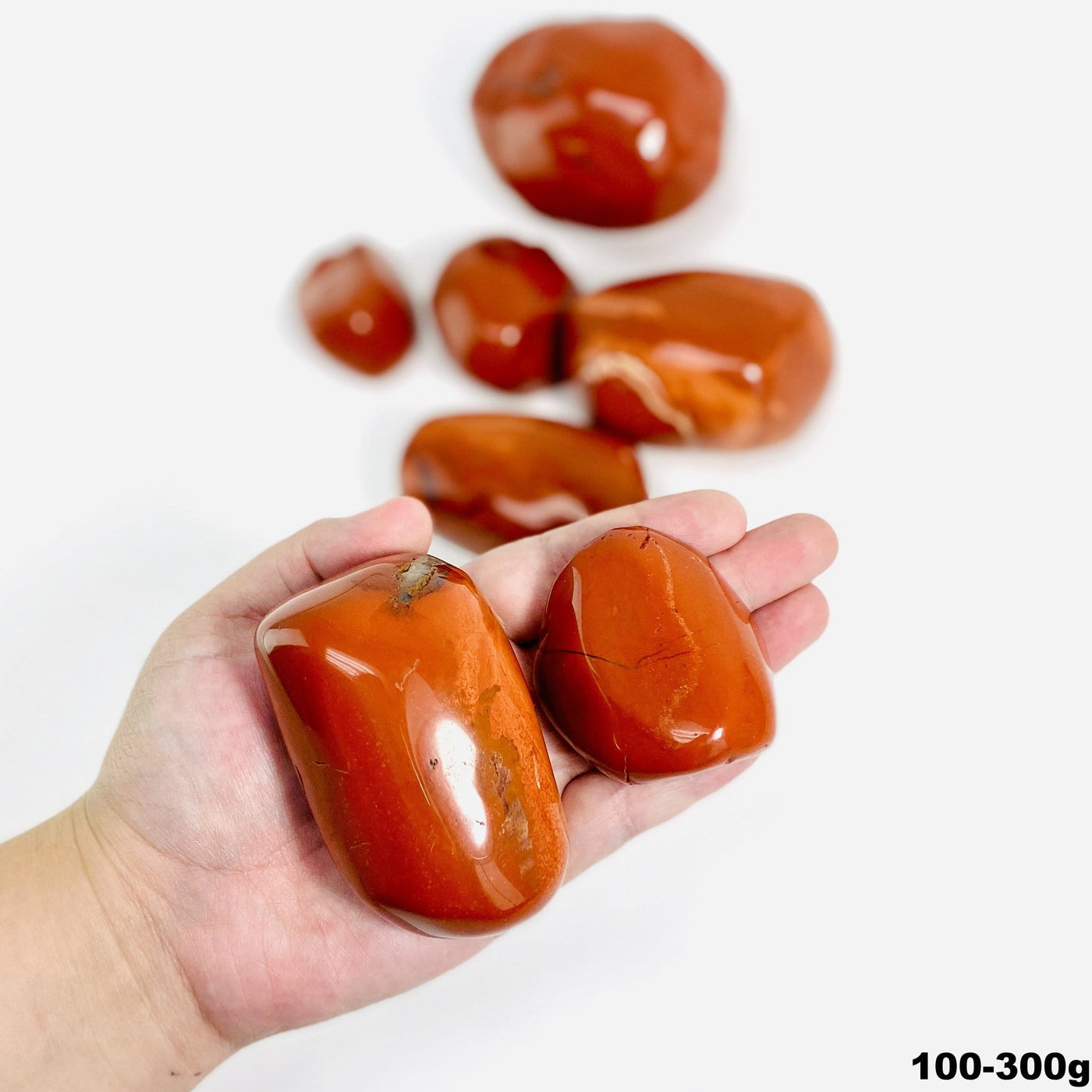Hand holding up 2 100-300g Red Jasper tumbled Stones with others blurred on white background