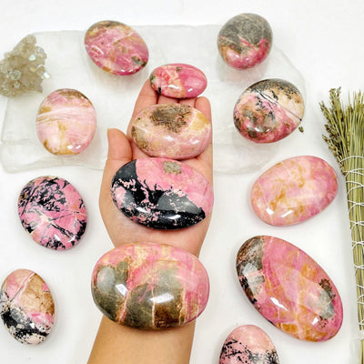 Hand holding up 4 Rhodonite Palm Stones with others and decorations in the background