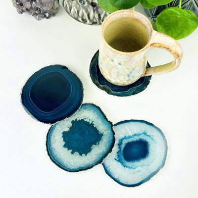 3 Teal Agate coasters with a silver electroplated edge next to 1 piece that is under a coffee cup for display, Slices measure about 3.5-5"