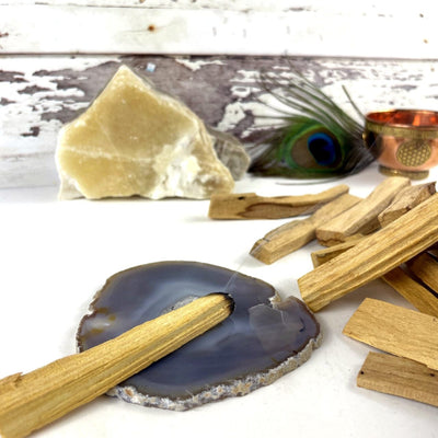 A pile of palo santo sticks on a white background.  One is lit and the tip is placed on a gray agate slice.