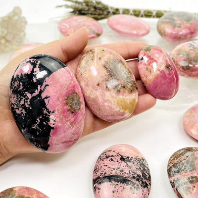 Hand holding up 3 Rhodonite Palm Stones with others and decorations blurred in the background