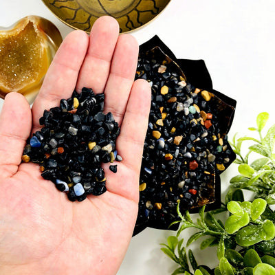 Black onyx chips in a lotus bowl.  They are mixed with a few other colorful stones. Some are held in a hand above the bowl.