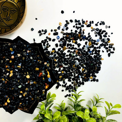Black onyx chips in a lotus bowl.  They are mixed with a few other colorful stones.  Some are spilled out of the bowl on a white background.
