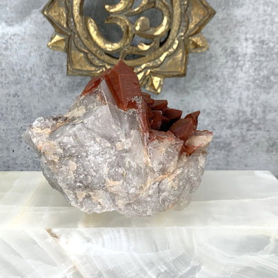 Close up photo of the back of the red quartz cluster, focusing on the clear quartz formations on the bottom of the cluster, red quartz cluster is also being displayed on a white and grey marbled back ground.