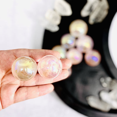 Hand holding up 2 Rose Quartz Angel Aura Titanium Spheres with others in a black container blurred in the background
