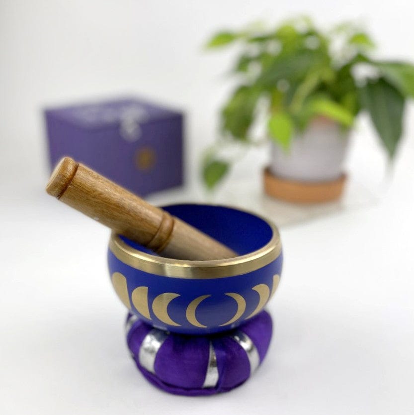 Purple singing bowl with moon phase design around sides, sitting atop a pillow, with the mallot sitting inside the bowl