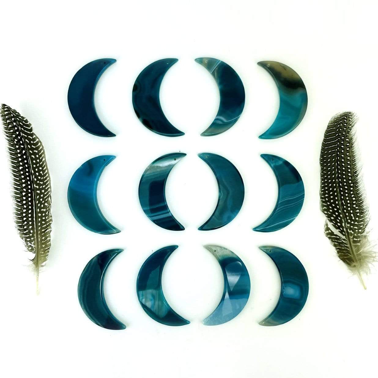 Twelve teal agate moons displayed on a white surface next to feathers.