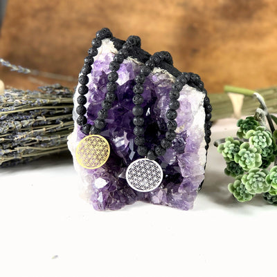 Diffuser Necklace Lava Bead with Flower of Life Charm in Gold/Silver tone in Top of Amethyst Cluster on Brown Background.