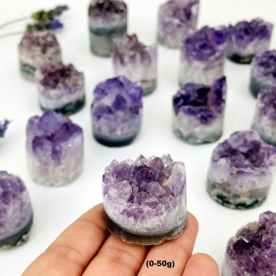 Amethyst cores with shades of white and purple in assorted sizes to show variations on a white background. 0-50g is in a woman's hand with others in the background.