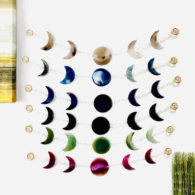 photo of all six colors of moon phase wall hanging on the wall.  They are natural tanish agate (can vary) blue, teal , black, green, and pink.  Some have agate banding some do not.  Crystal quartz beads between each moon phase.  Center round agate for the full moon and two cresent moons on each side