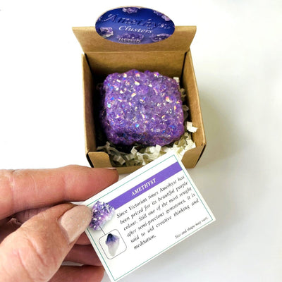 hand holding up card explaining amethyst aura geode in box with decorations in the background