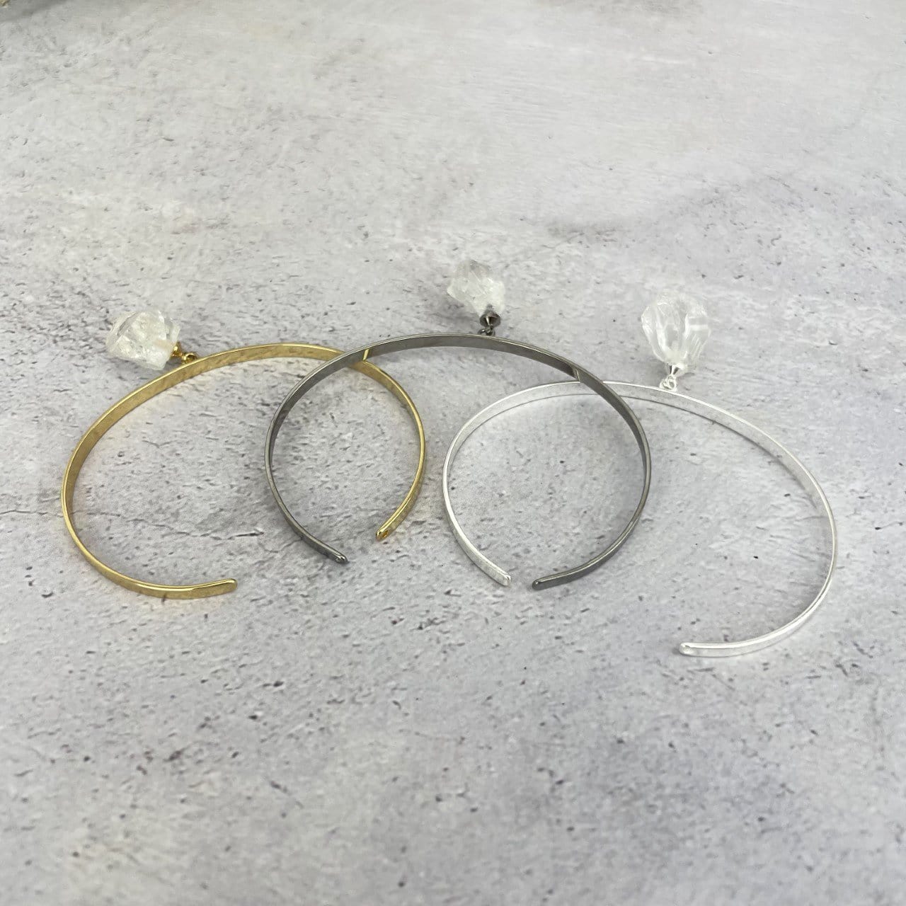 Gold and silver bracelets that are thin metal with a rough crystal quartz hanging from the middle.  Pictured here on a purple and white point. Pictured here in gold plated, gun metal plated, and silver plated on a cement background.