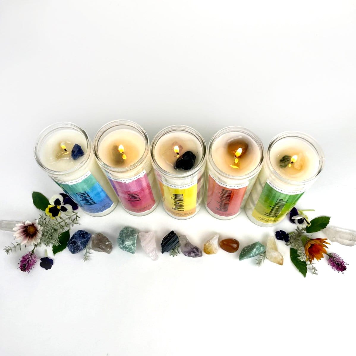 Different candles lined up surrounded by crystals and flowers on white background