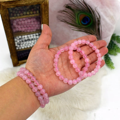 Hand wearing 2 Rose Quartz Round Bead Bracelets and holding 2 others with display full of bracelets blurred in the background
