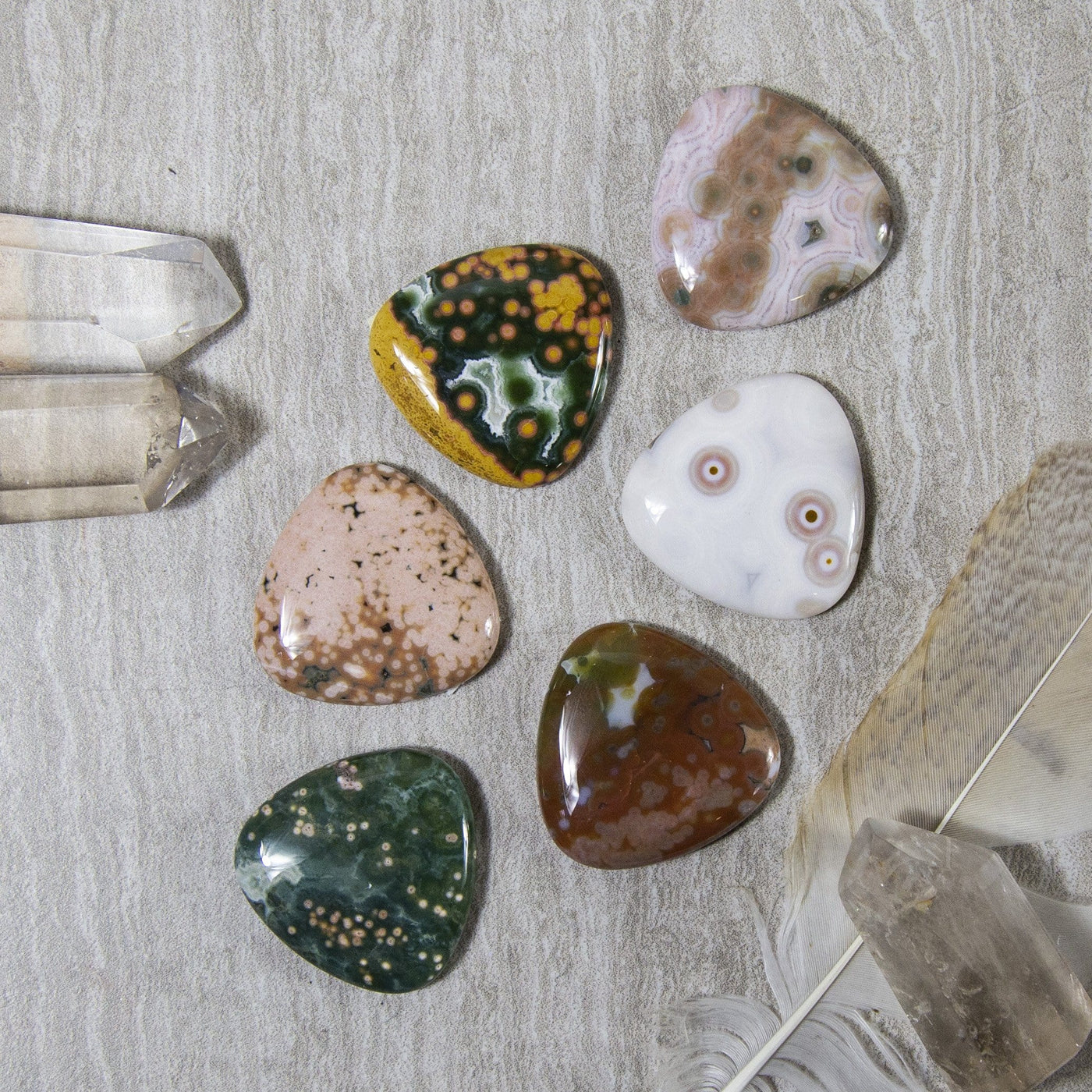 multiple Triangle Shaped Jasper Cabochons displayed on gray background showing various patterns and color formations such as spots waves of line formations greens browns yellows whites tan 