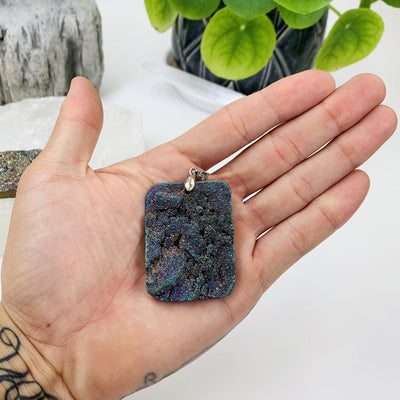 Close photo to the smallest pendant for this rainbow titanium pendant lot listing. The item is being held for size reference and its also being displayed on a White back ground in front of an all natural green plant.