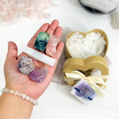 hand holding up fluorite, rose quartz, selenite lepidolite, and amethyst with serenity heart box in the background