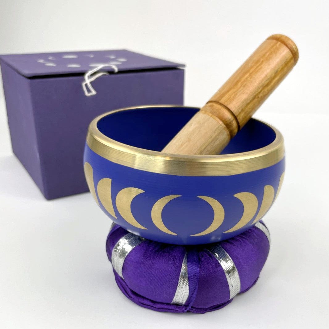 purple singing bowl with gold moon phase design sitting on top of pillow, with wood mallot sitting inside and purple box in the background