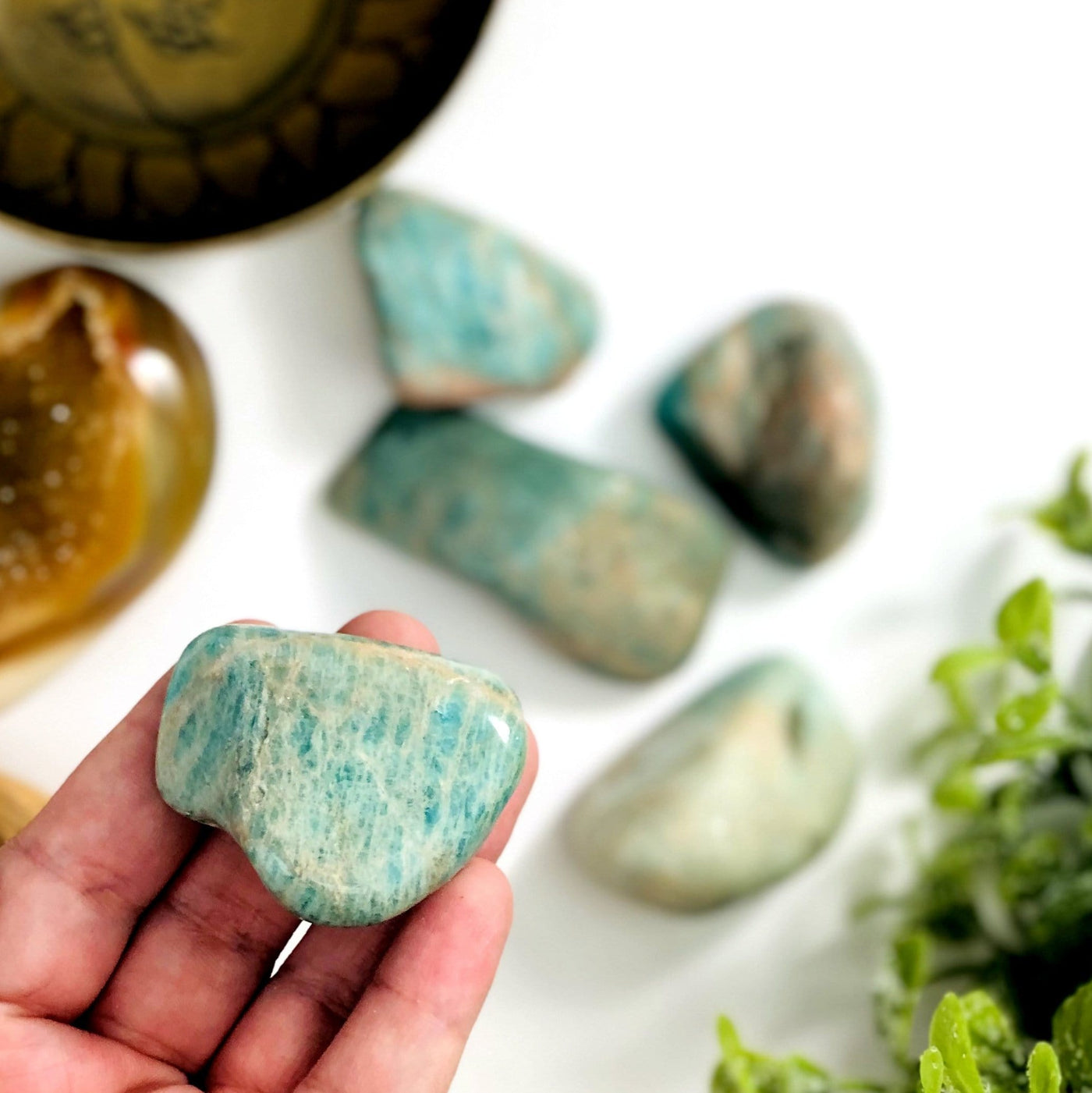 Amazonite tumbled stones.  They are a greenish blue shade and some have tan markings.  One is held in a hand and other are blurred on a white background.
