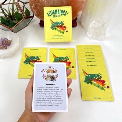 An affirmation card in a hand, card deck box and expanded cards within an alter.