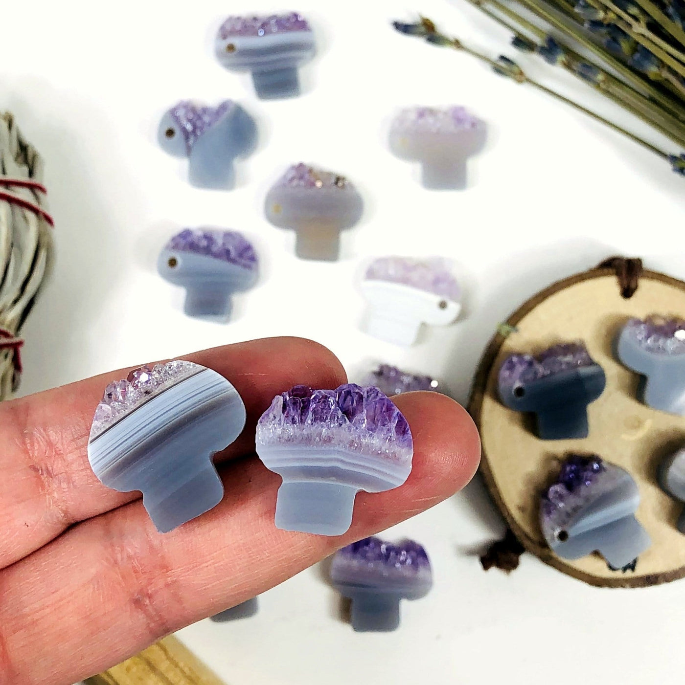 Amethyst slice cut into mushroom shapes with a drill hole on the side.  This photo shows an assortment as they range in colors and patterns with shades of white, gray, brown, purple and tan.  Two are held in a hand and the rest are in the background.