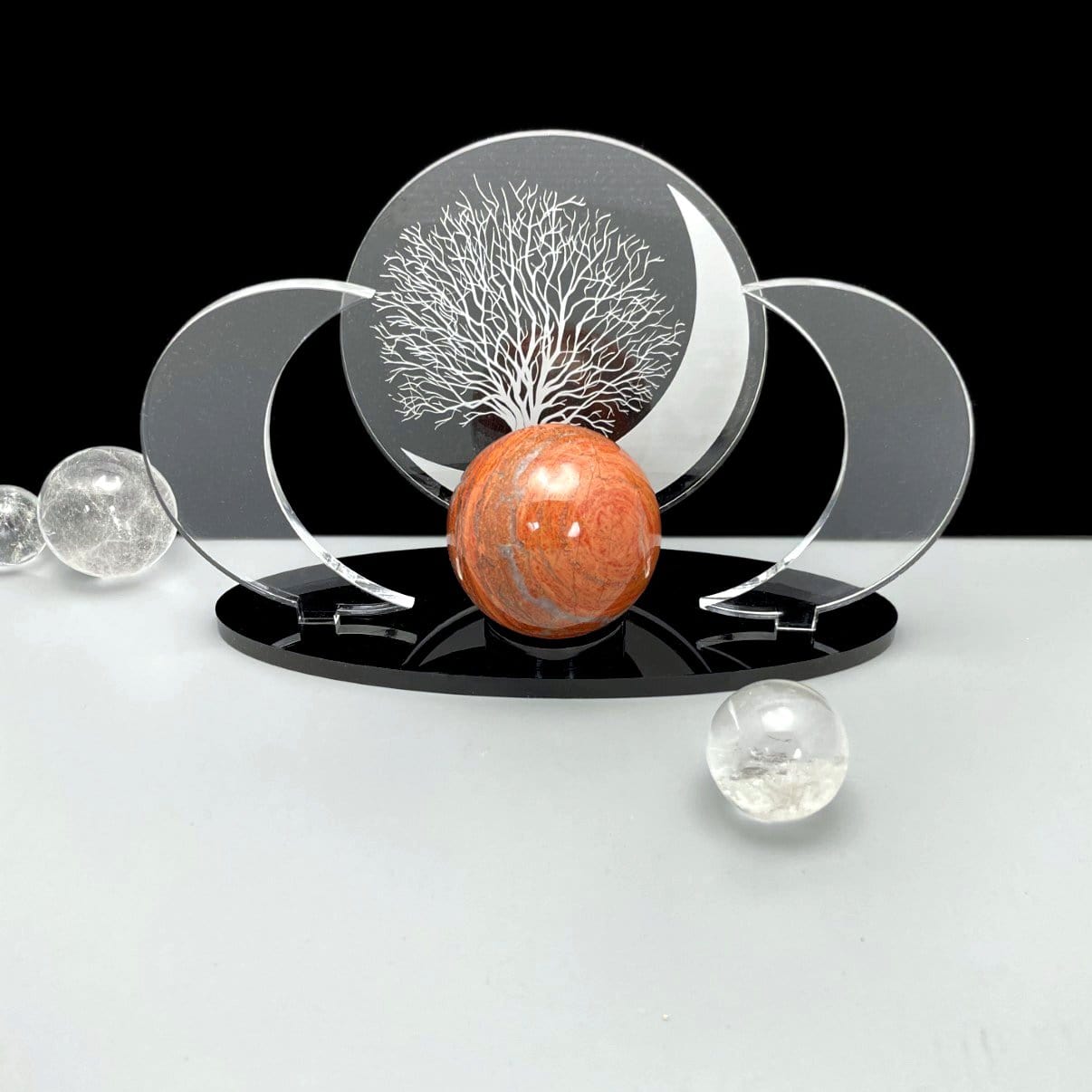 Sphere holder with a cut out acrylic circle in the middle that has an engraved tree and moon.  Two clear acrylic moons on the side and a black base.  In this photo a red jasper sphere is placed on it.