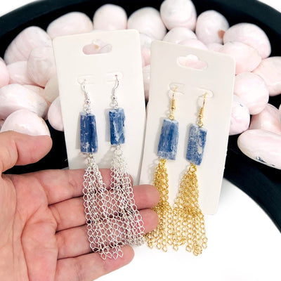 hand holding Blue Kyanite Dangle Earrings with Electroplated 24k Gold/Silver Tassels with decorations in the background