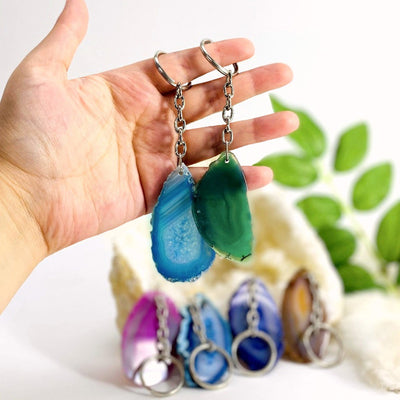 Two agate keychains in a hand. Multiple agates in the background with plants.