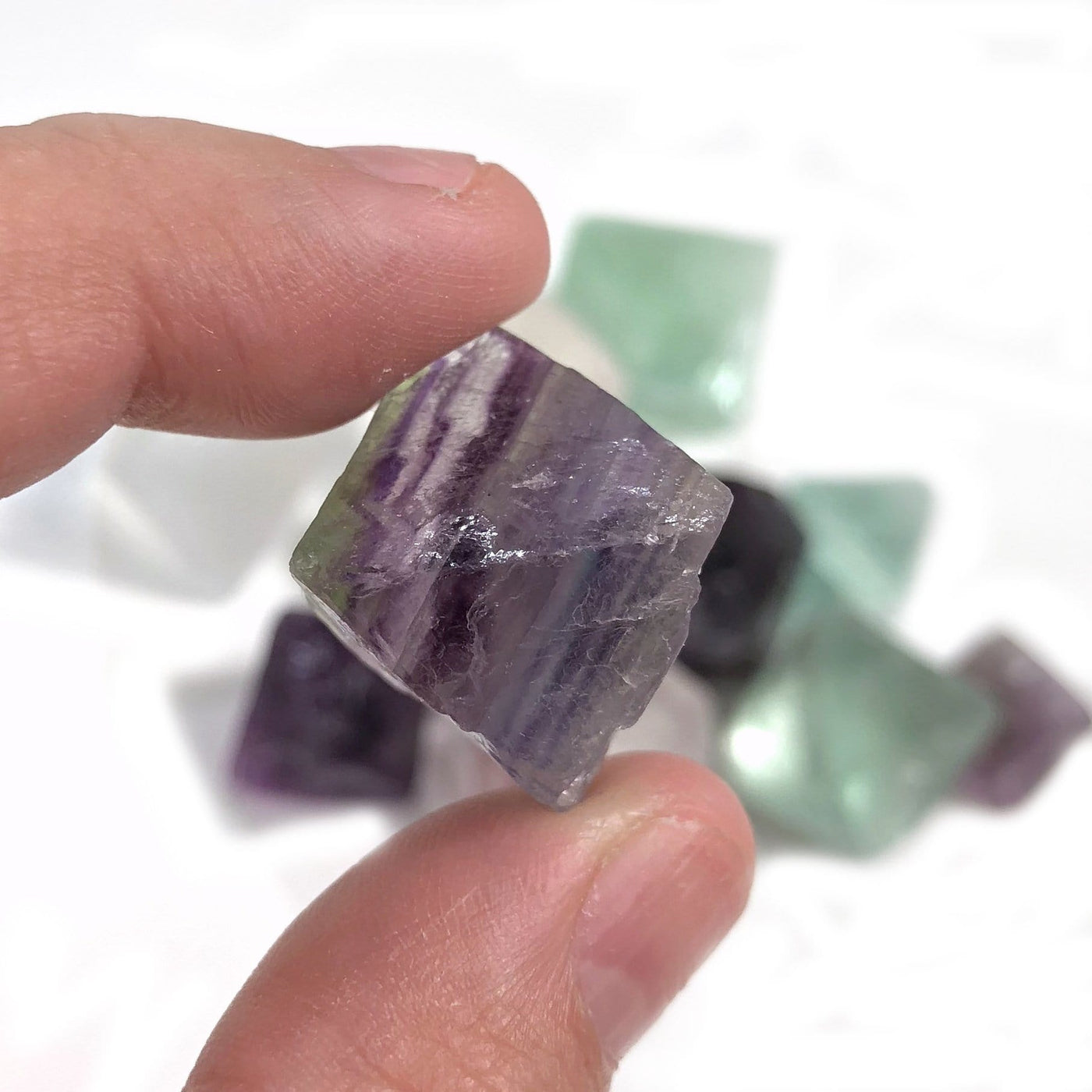 Fingers holding up 1 Rainbow Fluorite Octahedron Cube with other Rainbow Fluorite Octahedron Cubes blurred in the background