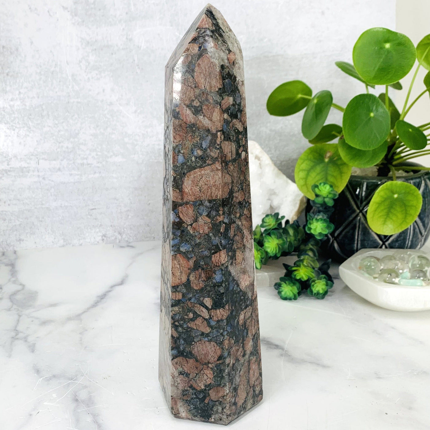 Side shot of Rhyolite Polished Tower, the Rhyolite tower is being displayed on a marbled background, next to a green natural plant.