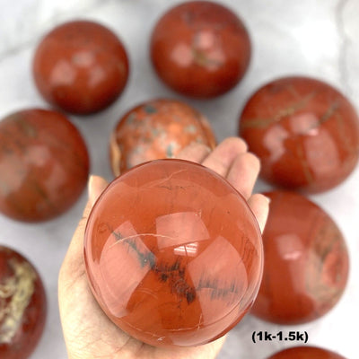 Hand holding up 1-1.5k Red Jasper Sphere  with others blurred on gray background