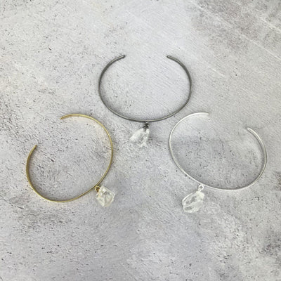 Gold and silver bracelets that are thin metal with a rough crystal quartz hanging from the middle.  Pictured here on a purple and white point. Pictured here in gold plated, gun metal plated, and silver plated on a cement background.