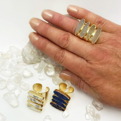 A hand wearing the Natural Druzy Adjustable Teardrop Druzy Ring with Electroplated 24k Gold Adjustable Cigar Band while Mystic Blue Titanium Druzy and White Howlite, are on a table underneath 