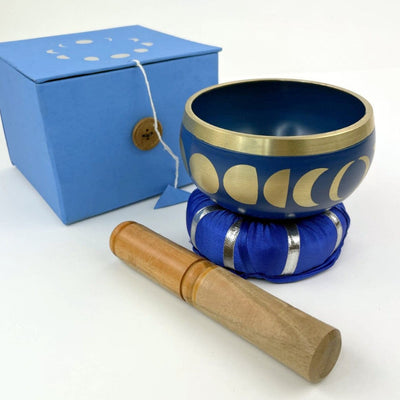 Blue singing bowl from the side showing moon phase design in gold sitting on top of pillow and mallot beside it and blue box behind