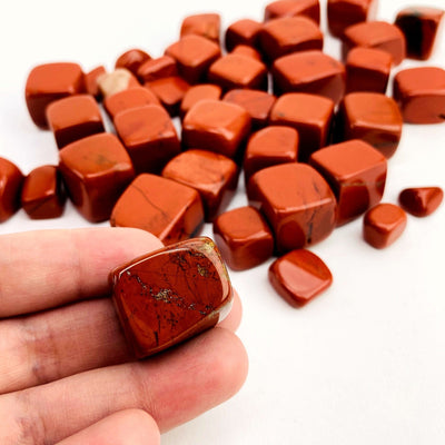 Hand holding up Red Jasper Cubed Tumbled Stone with others scattered on white background