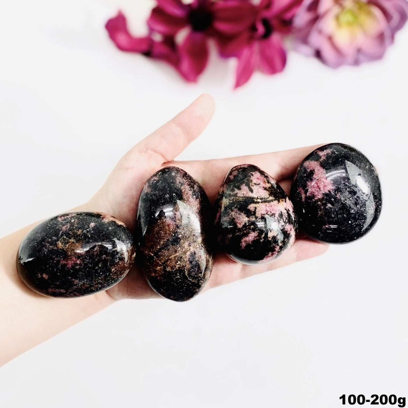 Hand holding up 100-200g Rhodonite Palm Stones with flowers blurred on white background