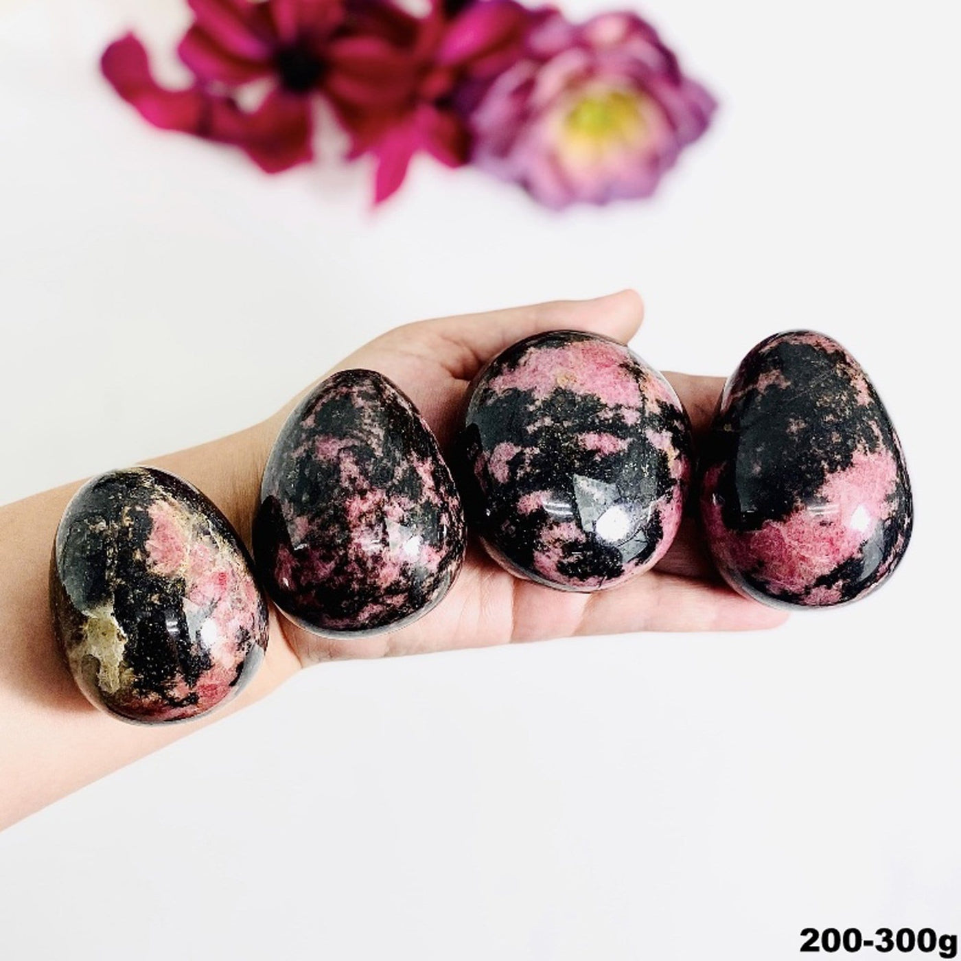 Hand holding up 200-300g Rhodonite Palm Stones with flowers blurred on white background