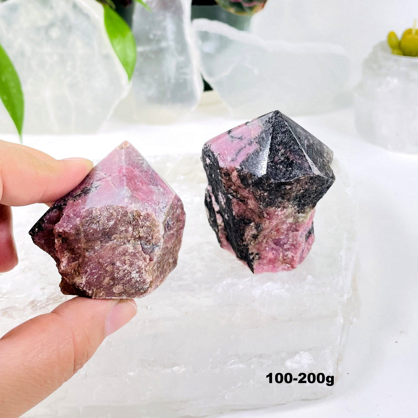 Hand holding 100-200g Rhodonite Point with another on a crystal quartz slab and decorations blurred in the background