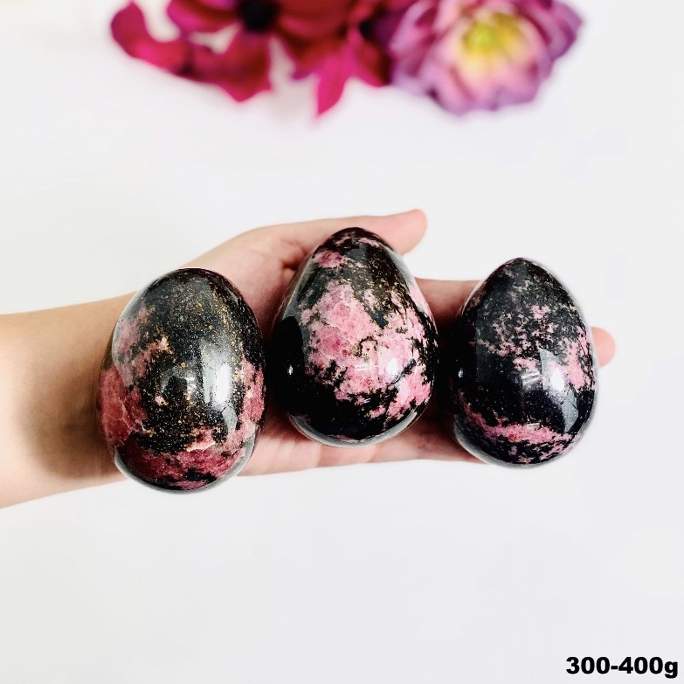 Hand holding up 300-400g Rhodonite Palm Stones with flowers blurred on white background