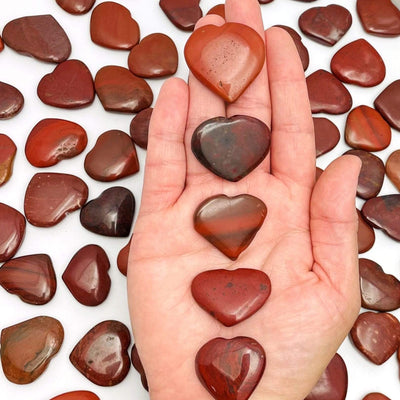 Hand holding up 5 Red Jasper Heart Shaped Stones in front of more scattered on white background
