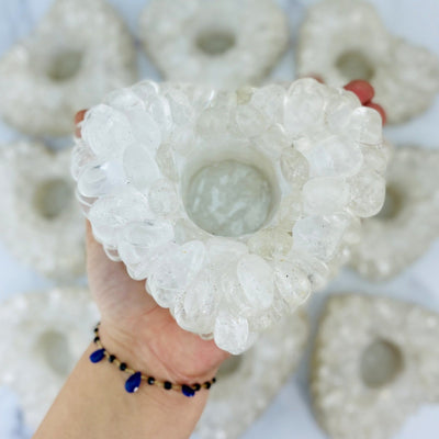 Heart shaped candle holder that is made with crystal quartz tumbled stones that are glued together.  Shown held in a woman's hand with more candle holders blurred in the background.
