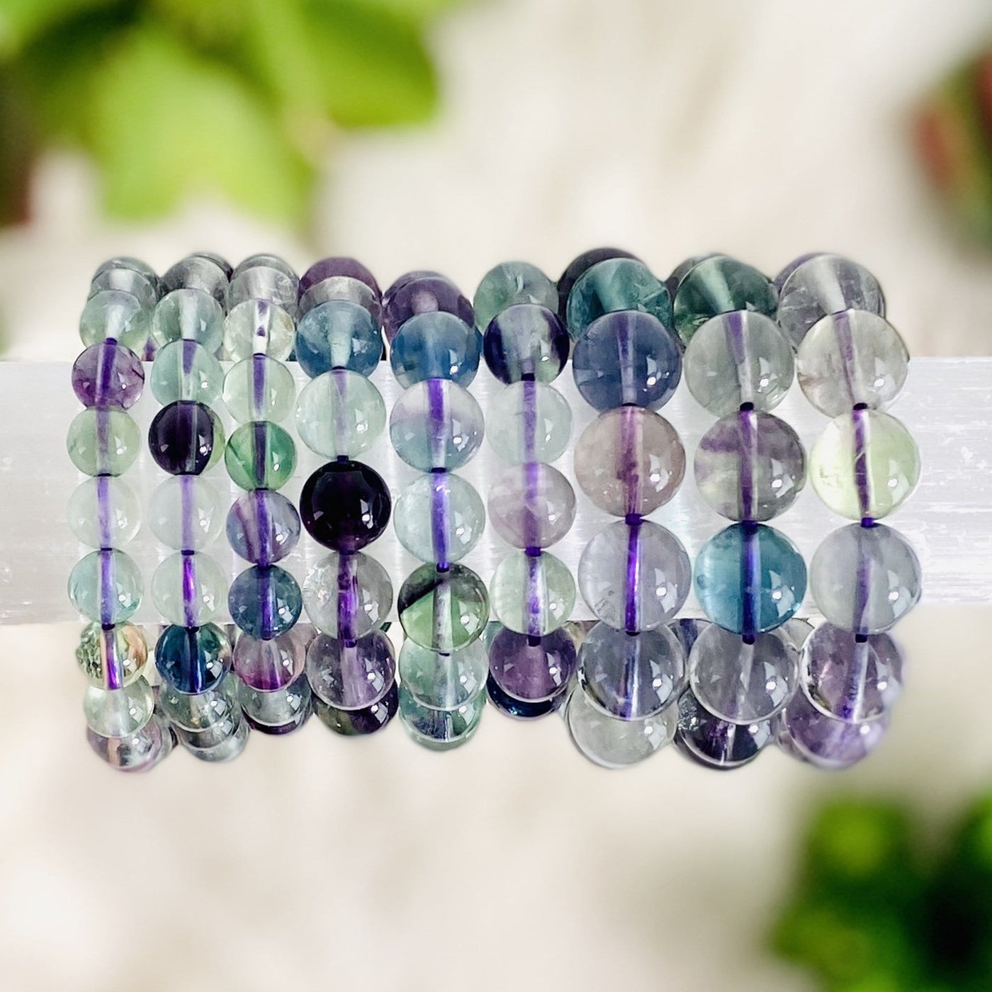 9 Rainbow Fluorite Round Bead Bracelets displayed on bracelet stand with plants blurred on white background
