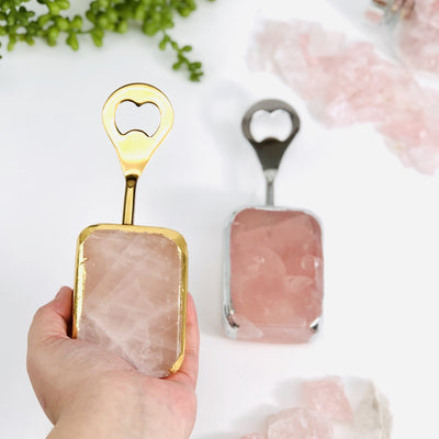 Hand holding up gold Rose Quartz Bottle Opener while silver bottle opener is in the background with crystals and plants