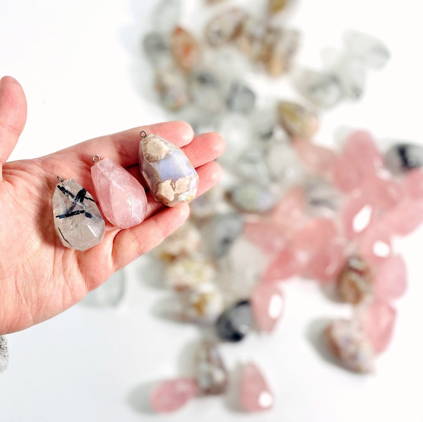Hand holding up 1 of each Rose Quartz Rutilated Quartz Flower Agate Pendant with pile of others blurred in the background