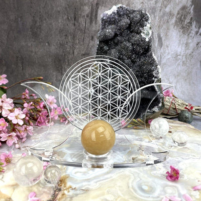 A front view of an Acrylic Sphere Holder Crescent Moons - Flower of Life holding a sphere in an alter that consists of crystals and flowers.