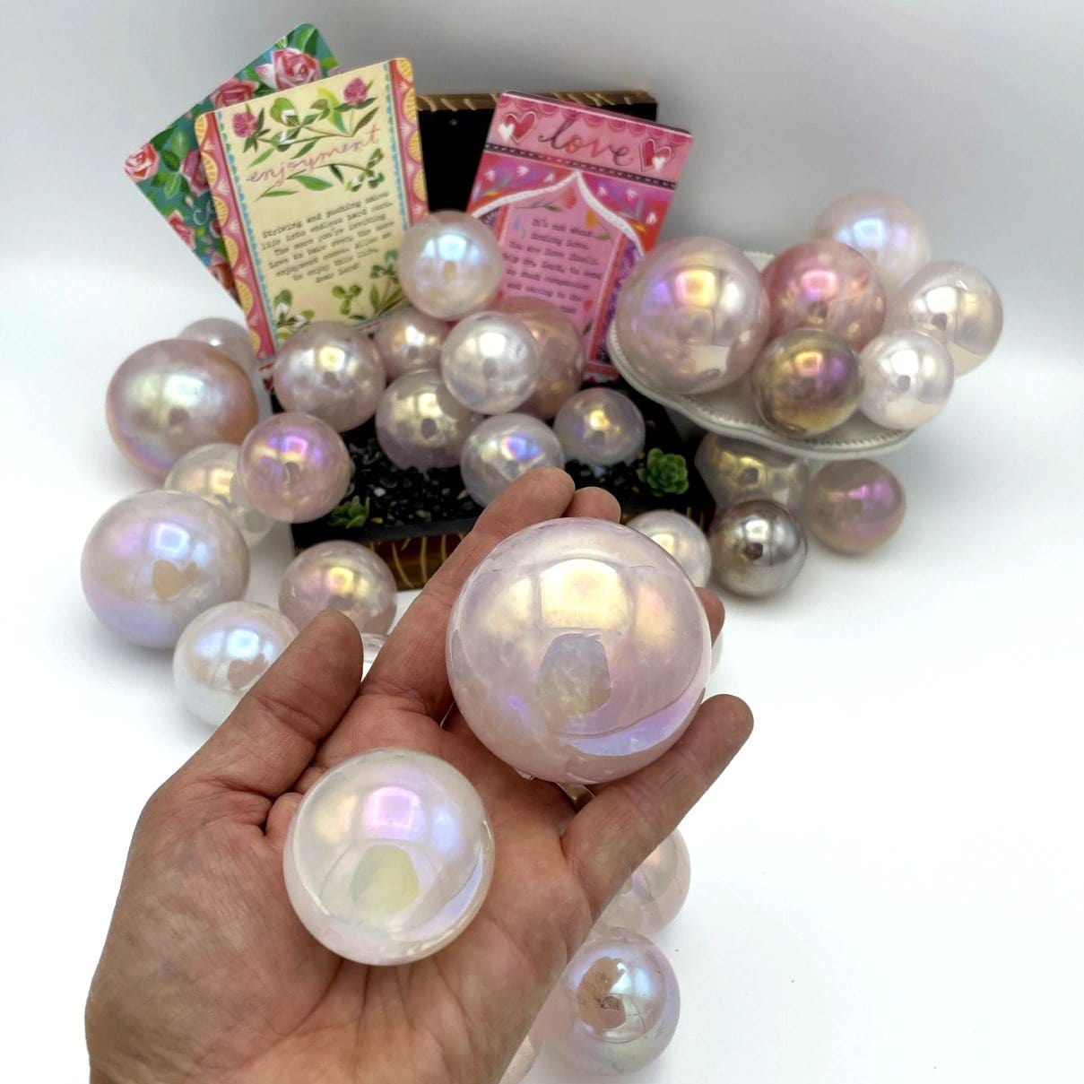 Hand holding 2 Rose Quartz Angel Aura Titanium Spheres of different sizes in front of more spheres and decorations in the background
