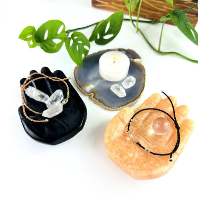 Hamsa Hand Stone Carvings in black obsidian with small crystals and a bracelet in the hand, and an orchid calcite with a bracelet and small sphere in the palm