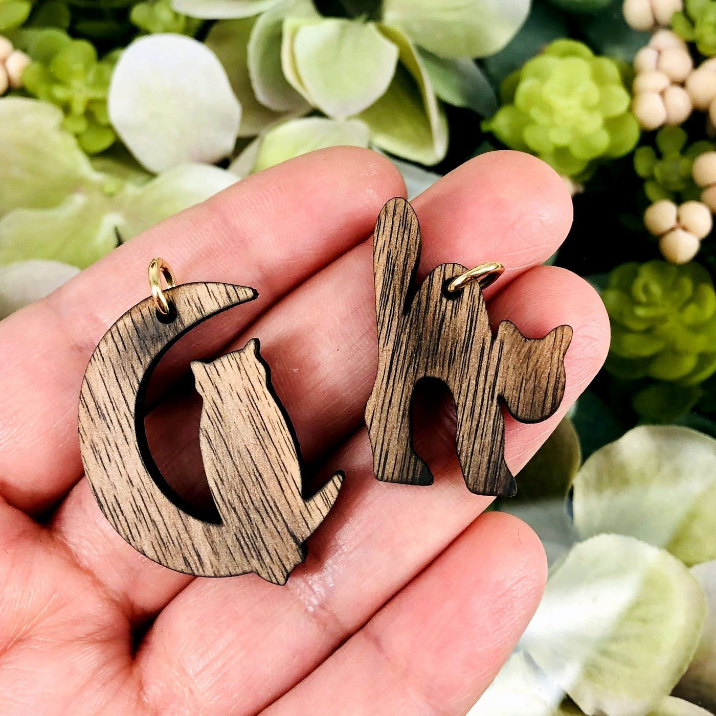 hand holding up 2 wooden cat pendants with decorations in the background