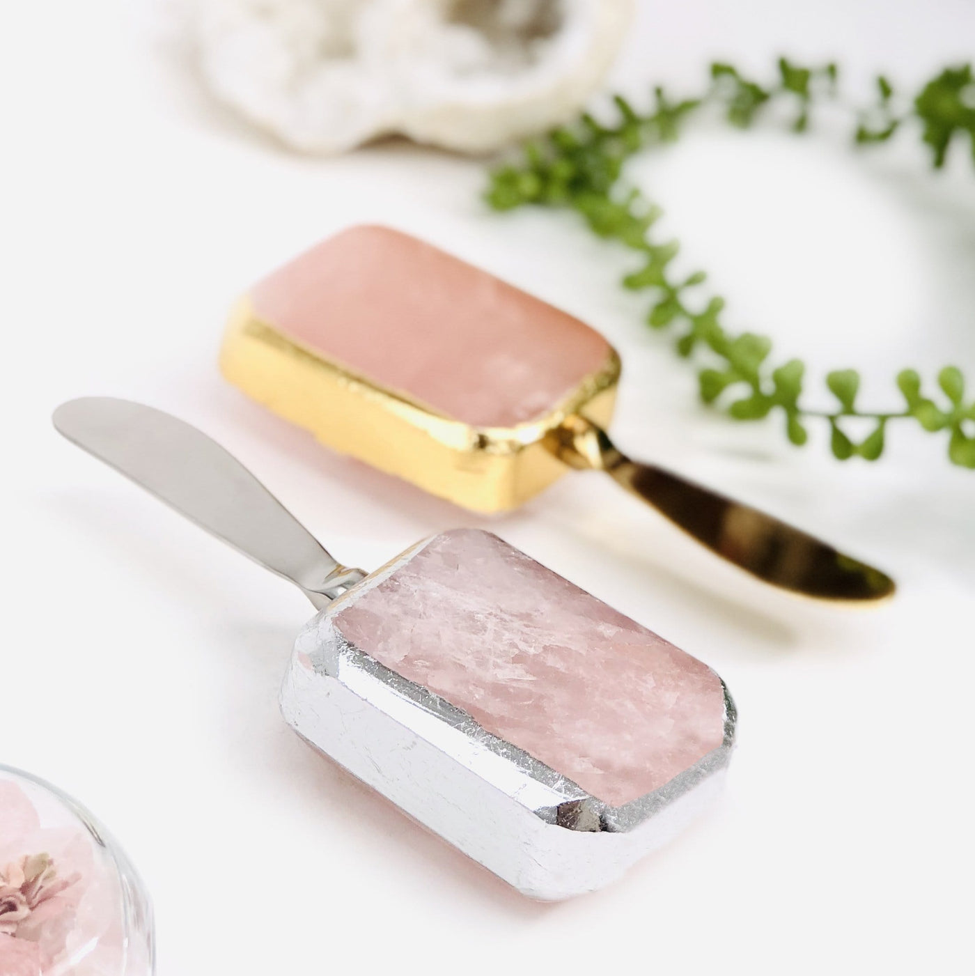 Up close shot of 2 Rose Quartz Butter Spreaders with plants blurred in the background